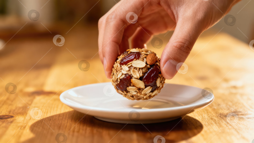 Скачать Рука, берущая с блюдца самодельный энергетический шарик. Полезный веганский перекус с овсяными хлопьями, миндалем и сухофруктами на деревянном столе фотосток Ozero