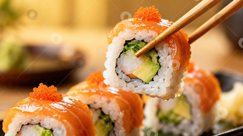 Скачать Крупный план суши-ролла с лососем, который держат палочками для еды. Японская кухня со свежей рыбой, авокадо и икрой тобико. Вкусные блюда азиатской кухни и блюда из морепродуктов фотосток Ozero
