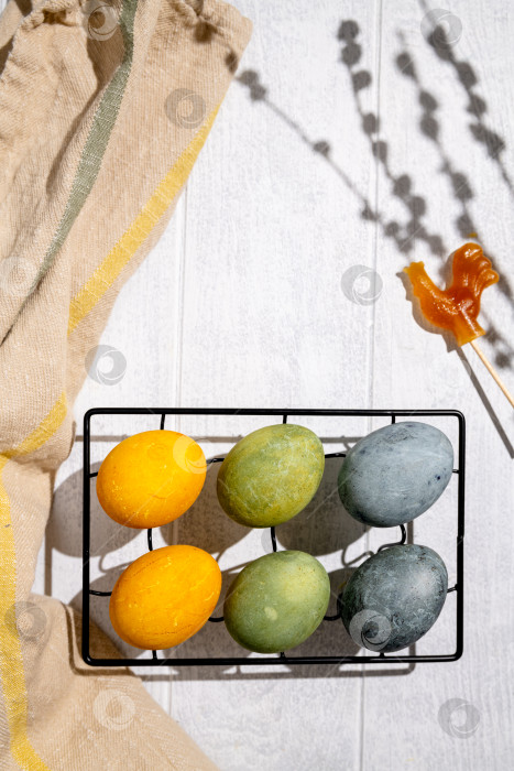 Скачать Пасхальные яйца, окрашенные натуральным способом с добавлением гибискуса и куркумы, лежат на подставке для охлаждения на белом деревянном столе, окруженные этикетками, веточками вербы, бечевкой и ножницами фотосток Ozero