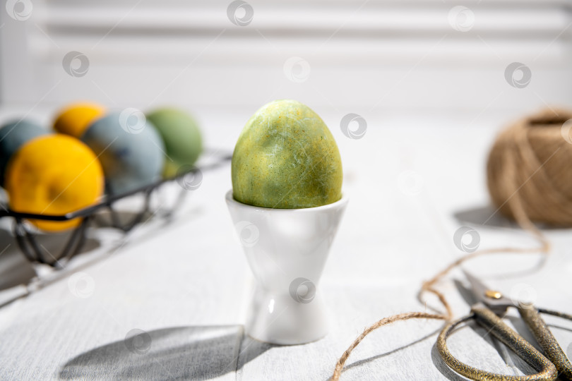 Скачать Зеленое пасхальное яйцо стоит в белой чашке для яиц на белом деревянном столе вместе с другими разноцветными яйцами в черной металлической подставке для яиц, шпагатом и ножницами, создавая праздничную пасхальную сцену. фотосток Ozero
