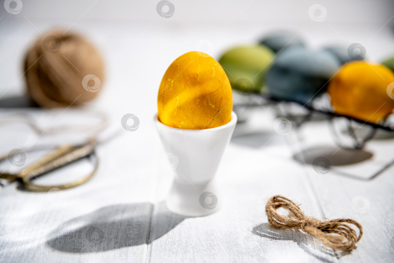 Скачать желтое пасхальное яйцо лежит в белой чашке для яиц на деревянном столе, окруженное принадлежностями для рукоделия и разноцветными размытыми яйцами, создавая праздничную атмосферу фотосток Ozero