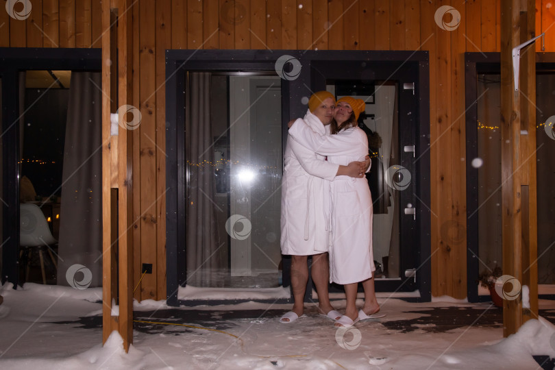 Скачать Пара отдыхает на террасе загородного дома зимним вечером. Красивая женщина и красивый мужчина расслабленно стоят на террасе деревянного коттеджа. Зимний курорт. фотосток Ozero