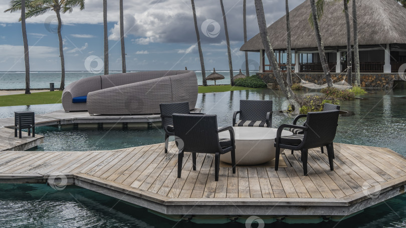 Скачать На дощатых террасах у бассейна установлены стол, стулья и полукруглый диван из плетеного ротанга. фотосток Ozero