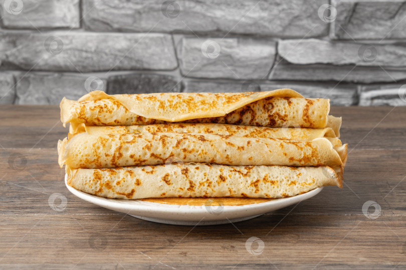 Скачать Стопка тонких блинчиков на белой тарелке, поставленная на деревянный стол у каменной стены на кухне в светлое время суток фотосток Ozero