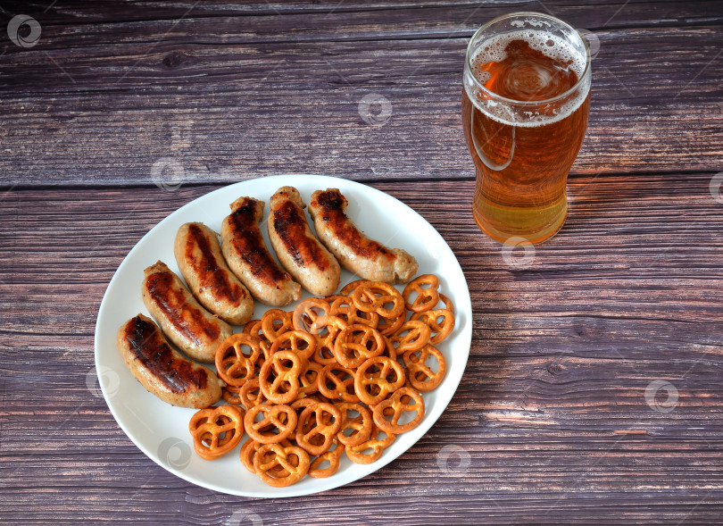 Скачать Стакан светлого пива и тарелка с жареными сосисками и солеными оладьями на деревянном столе. фотосток Ozero