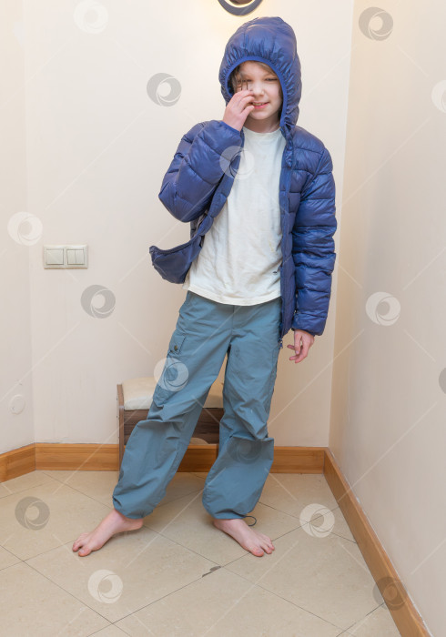 Скачать Маленький мальчик в синей пуховой куртке с поднятым капюшоном. Стоит в помещении, улыбается и дотрагивается до своего носа. Место для копирования фотосток Ozero