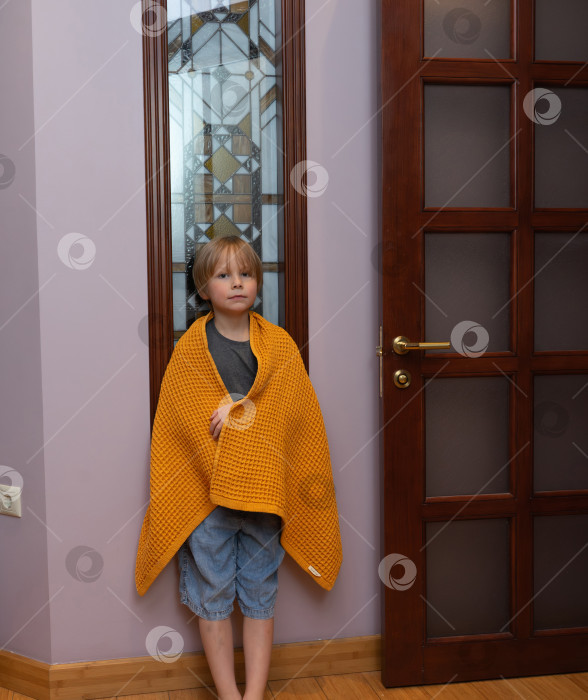 Скачать Маленький мальчик, завернутый в желтое одеяло, стоит у двери. Ребенку уютно и тепло фотосток Ozero