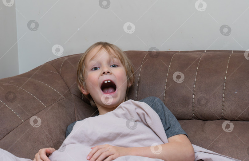 Скачать Уставший мальчик, зевающий с широко открытым ртом на диване. Сонный ребенок просыпается дома. фотосток Ozero