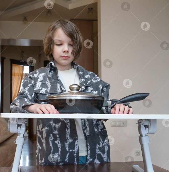 Скачать Маленький мальчик стоит со сковородкой на столе. Ребенок ждет ужина на кухне фотосток Ozero