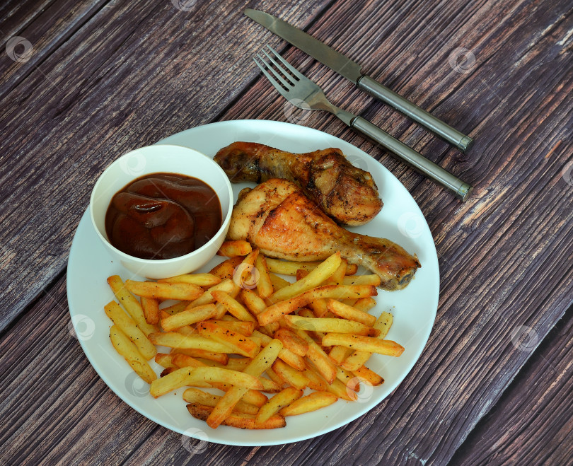 Скачать Запеченные куриные ножки с картофелем фри и кетчупом в круглой тарелке на деревянном столе, рядом со столовыми приборами. фотосток Ozero