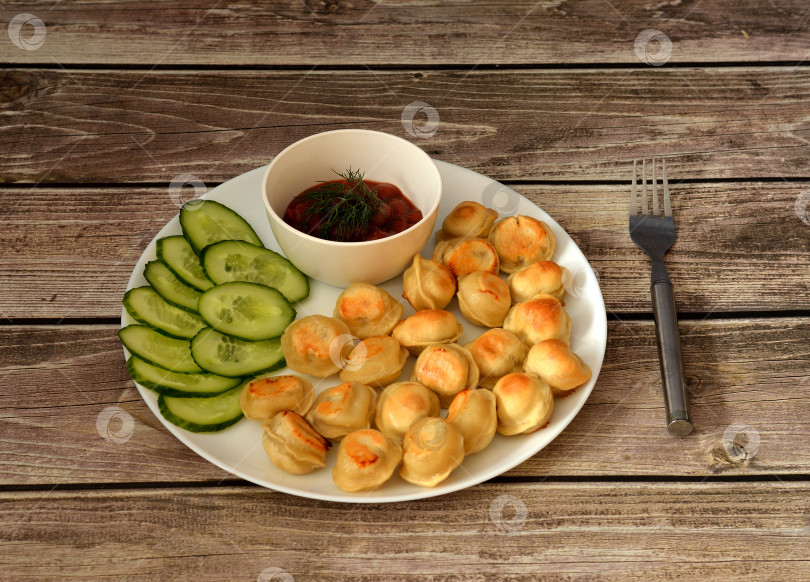Скачать Жареные клецки, нарезанный свежий огурец с чашечкой острого соуса в круглой белой керамической тарелке на деревянном столе, вилка рядом. фотосток Ozero