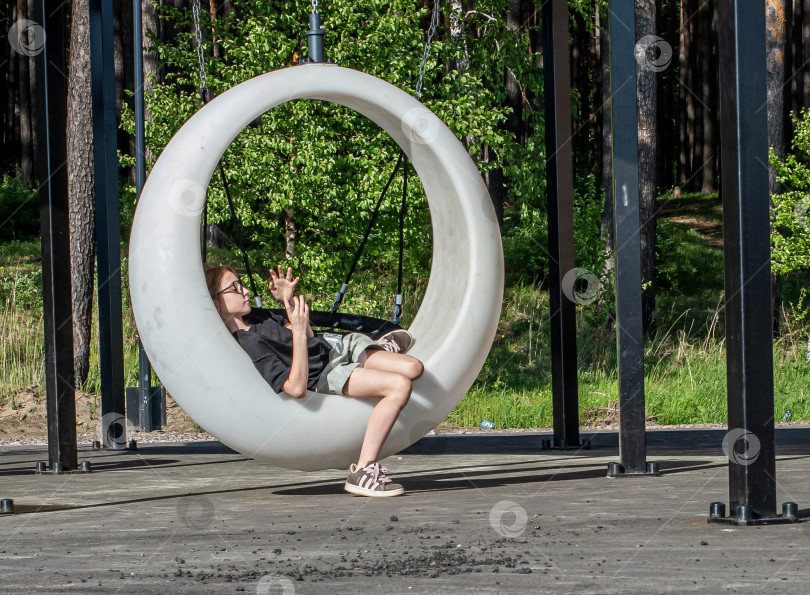 Скачать Россия,город Железногорск Красноярский Край, 25 мая 2025 год.Девочка качается на подвесных круглых качелях под навесом. Вокруг деревья и зелень. фотосток Ozero