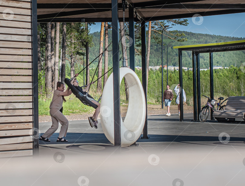 Скачать Россия,город Железногорск Красноярский Край, 25 мая 2025 год. девочка качается в подвесных круглых качелях под навесом. Вокруг деревья и зелень. фотосток Ozero