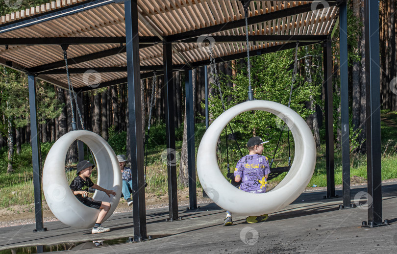 Скачать Россия,город Железногорск Красноярский Край, 25 мая 2025 год.Два мальчика сидят в подвесных круглых качелях под навесом. Вокруг деревья и зелень. фотосток Ozero