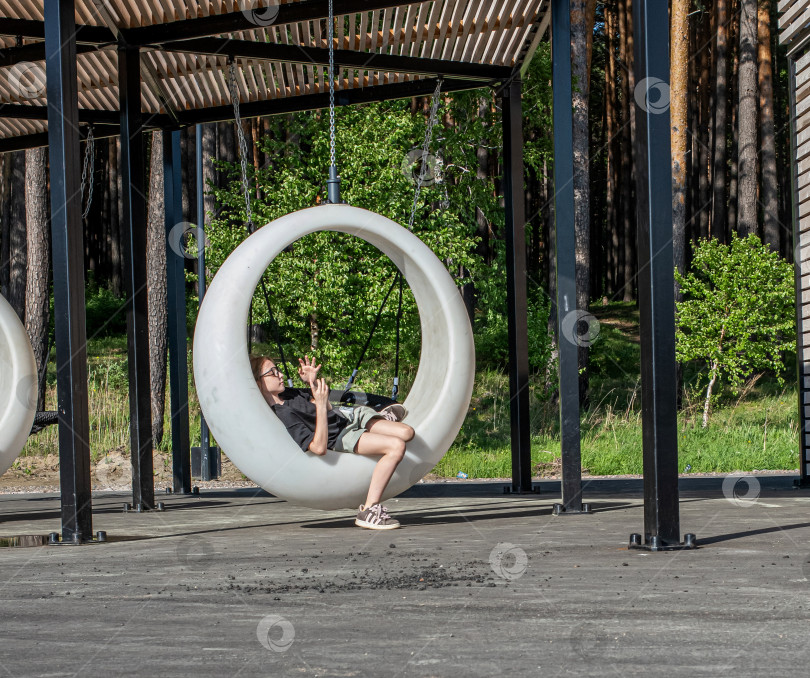 Скачать Россия,город Железногорск Красноярский Край, 25 мая 2025 год.Девочка качается на подвесных круглых качелях под навесом. Вокруг деревья и зелень. фотосток Ozero