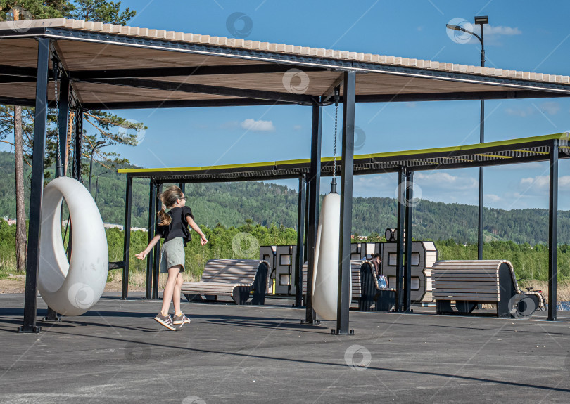 Скачать Россия,город Железногорск Красноярский Край, 25 мая 2025 год.Девочка на игровой площадке с качелями. Вокруг деревья и зелень. фотосток Ozero