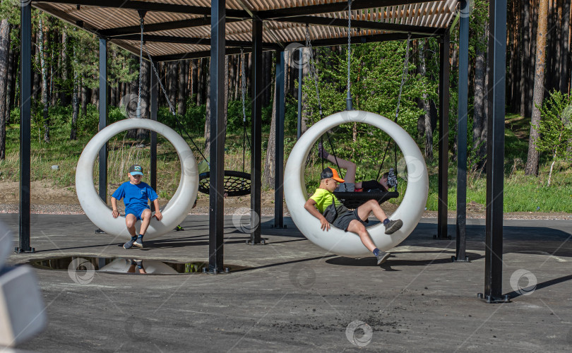 Скачать Россия,город Железногорск Красноярский Край, 25 мая 2025 год.Два мальчика сидят в подвесных круглых качелях под навесом. Вокруг деревья и зелень. фотосток Ozero