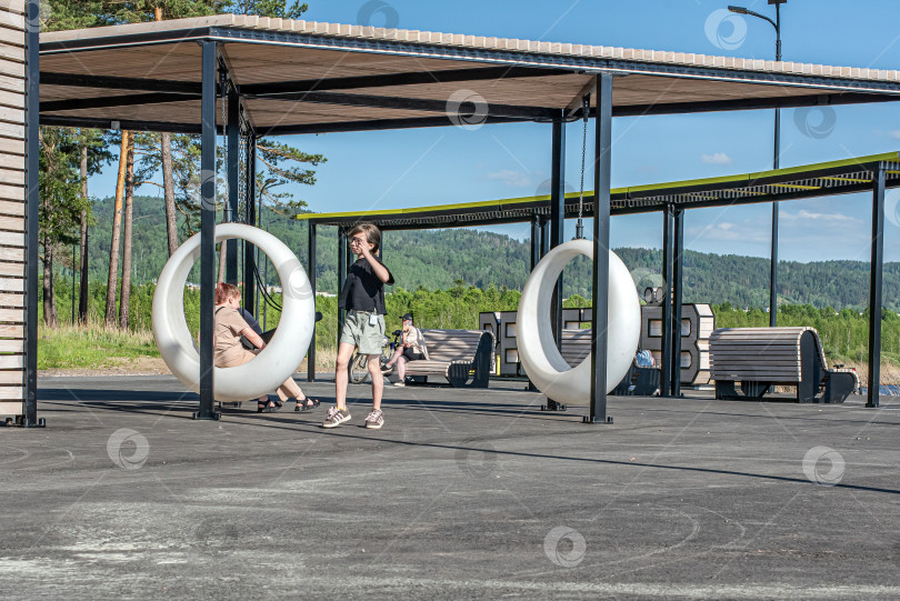 Скачать Россия,город Железногорск Красноярский Край, 25 мая 2025 год.Девочка качается на подвесных круглых качелях под навесом. Вокруг деревья и зелень. фотосток Ozero