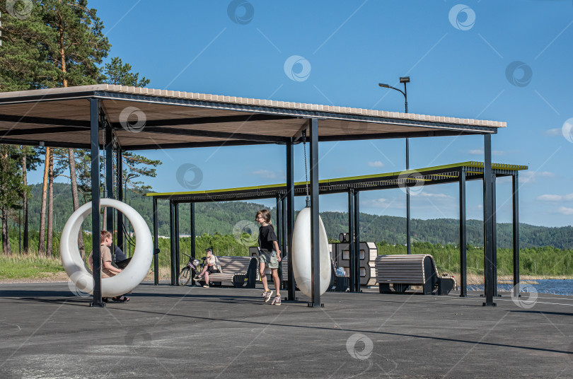 Скачать Россия,город Железногорск Красноярский Край, 25 мая 2025 год.Девочка качается на подвесных круглых качелях под навесом. Вокруг деревья и зелень. фотосток Ozero