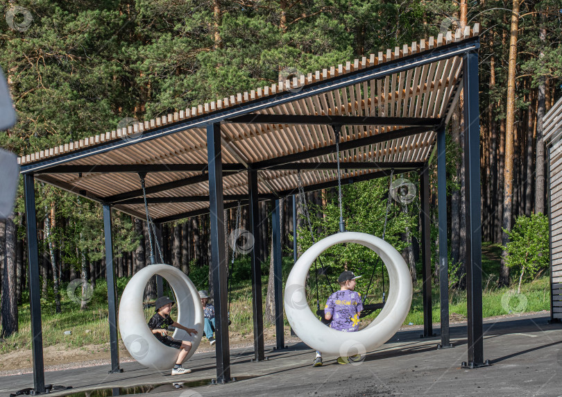 Скачать Россия,город Железногорск Красноярский Край, 25 мая 2025 год.Два мальчика сидят в подвесных круглых качелях под навесом. Вокруг деревья и зелень. фотосток Ozero