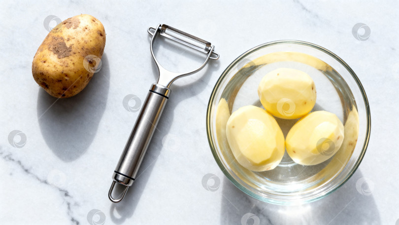 Скачать Целый картофель  и очищенный с картофелечисткой и миской  на мраморном фоне фотосток Ozero