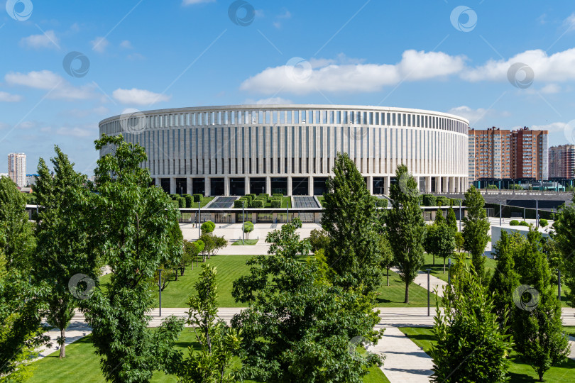 Скачать Многоуровневый ландшафтный городской парк "Краснодар" или "Парк Галицкого" на фоне здания футбольного стадиона "Краснодар". На переднем плане декоративные деревья. Краснодар, Россия - 18 июня 2021 года фотосток Ozero