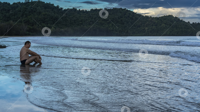 Скачать Раннее утро. Рассвет на берегу океана. Мужчина сидит в полосе прибоя, положив руки на колени. фотосток Ozero