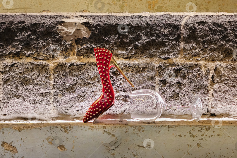 Скачать Красная туфля на высоком каблуке стоит на полке рядом с бокалом для вина. фотосток Ozero