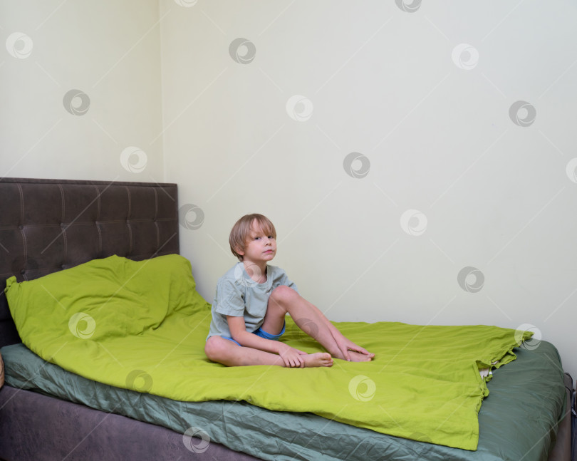 Скачать Маленький мальчик, сидящий на кровати с зеленым постельным бельем. Портрет светловолосого ребенка в спальне. Концепция домашнего образа жизни с пространством для копирования фотосток Ozero