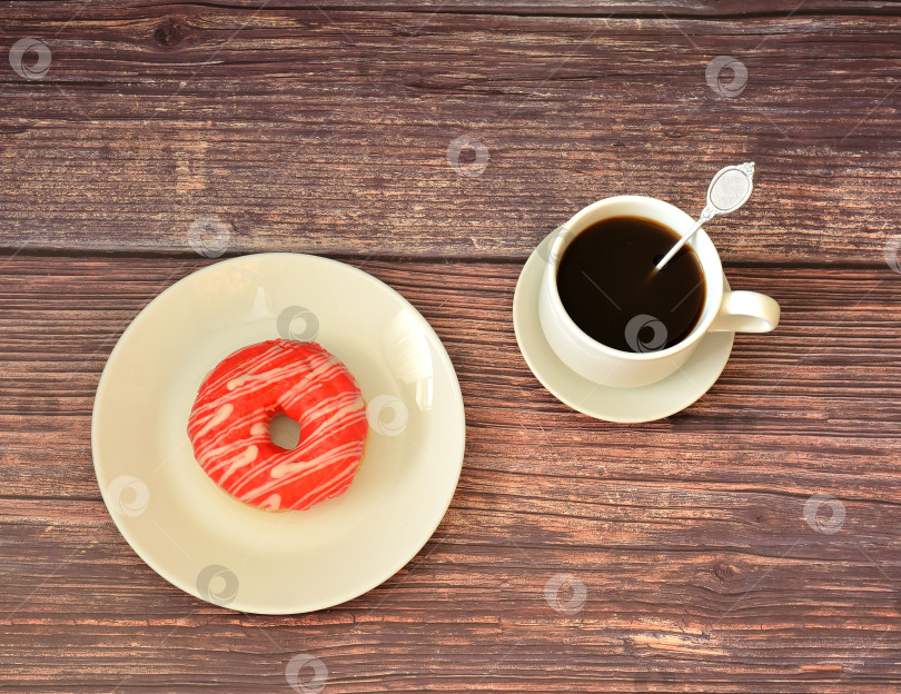 Скачать Свежий пончик в розовой ягодной глазури и чашка черного кофе на деревянном столе. фотосток Ozero