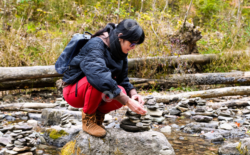 Скачать Пятидесятилетний русский путешественник-одиночка, сидящий на корточках у ручья и складывающий камни в осеннем лесу России, наслаждающийся тишиной, независимостью и осознанным путешествием в одиночку фотосток Ozero