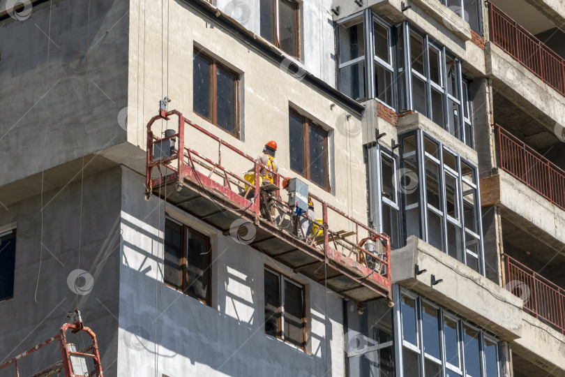 Скачать 21 августа 2025 года: Строительный подъемник с рабочими, выполняющими отделку стен на строительной площадке в Новороссийске, Россия, иллюстрирует современное строительство, рабочую силу и городское развитие. фотосток Ozero