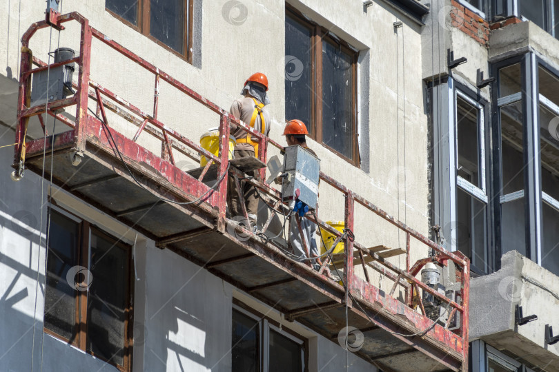 Скачать 21 августа 2025 года: Строительный подъемник с рабочими, выполняющими отделку стен на строительной площадке в Новороссийске, Россия, иллюстрирует современное строительство, рабочую силу и городское развитие. фотосток Ozero