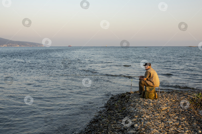 Скачать 24 августа 2025 года: Рыбак, сидящий на краю Суджукской косы, Россия, на фоне моря и кораблей, виднеющихся на горизонте, иллюстрирующих прибрежную жизнь, рыбную ловлю и морские пейзажи. фотосток Ozero