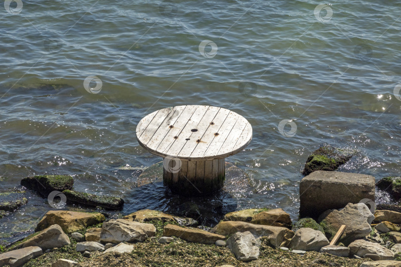 Скачать Большая деревянная катушка кабеля, лежащая на мелководье у берега, символизирует загрязнение окружающей среды, прибрежные отходы и экологические проблемы, затрагивающие морские районы и береговую линию. фотосток Ozero