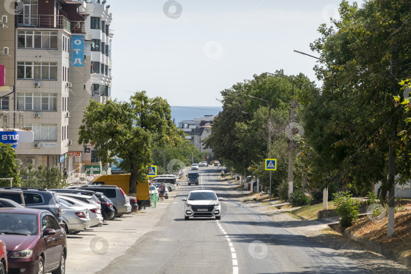 Скачать 24 августа 2025 года: Городская дорога в Мысхако, Россия, с автомобилями и жилыми домами, горами и морем на заднем плане, демонстрирующими повседневную жизнь и прибрежный городской пейзаж. фотосток Ozero