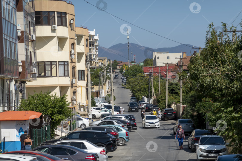 Скачать 24 августа 2025 года: Городская дорога в Мысхако, Россия, с автомобилями и жилыми домами, горами и морем на заднем плане, демонстрирующими повседневную жизнь и прибрежный городской пейзаж. фотосток Ozero