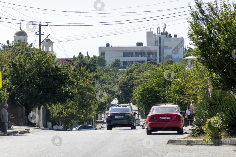 Скачать 24 августа 2025 года: Городская дорога в Мысхако, Россия, с автомобилями и жилыми домами, горами и морем на заднем плане, демонстрирующими повседневную жизнь и прибрежный городской пейзаж. фотосток Ozero