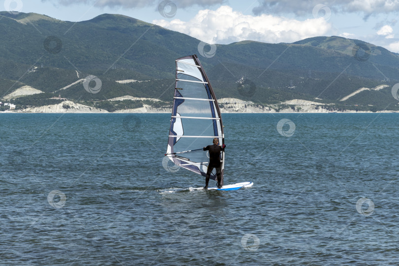 Скачать 24 августа 2025 года: Спортсмен занимается виндсерфингом в море недалеко от Новороссийска, Россия, с парусной доской и волнами, демонстрируя водные виды спорта, активный образ жизни и отдых на побережье. фотосток Ozero