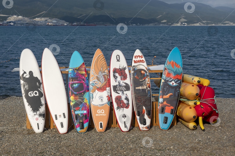 Скачать 28 июля 2025 года: Разноцветные доски для гребли, стоящие в ряд и сушащиеся на солнце на пляже в Новороссийске, Россия, отражают культуру летнего отдыха и водных видов спорта на Черноморском побережье. фотосток Ozero