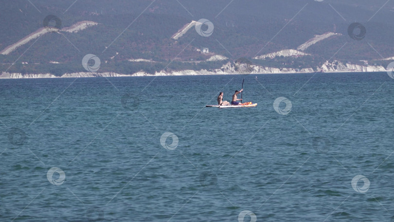 Скачать 23 августа 2025 года, Россия, Новороссийск. Мужчина и женщина плывут по морю на SUP-доске, наслаждаясь летними приключениями и живописными видами побережья. фотосток Ozero