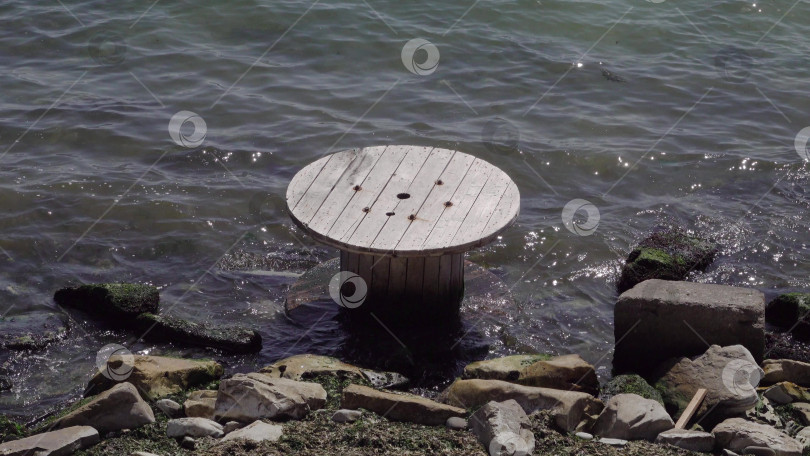 Скачать Большая деревянная катушка кабеля, лежащая на мелководье у берега, символизирует загрязнение окружающей среды, прибрежные отходы и экологические проблемы, затрагивающие морские районы и береговую линию. фотосток Ozero
