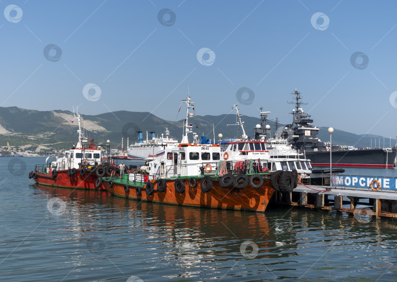 Скачать 12 августа 2025 года: Красные рабочие катера пришвартовались в оживленном порту Новороссийска, Россия, на фоне портовой деятельности и промышленной инфраструктуры. фотосток Ozero