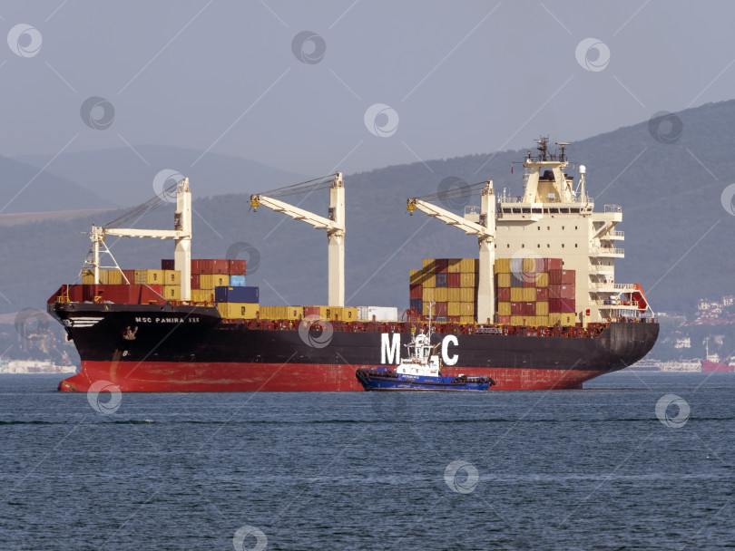 Скачать 12 августа 2025 года: Грузовое судно MSC Pamira III заходит в порт Новороссийск, Россия, двигаясь по спокойным водам на фоне оживленной портовой жизни. фотосток Ozero