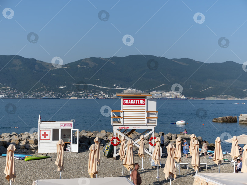 Скачать 12 августа 2025 года: Городской пляж в Новороссийске, Россия, с медицинским пунктом и спасательной вышкой, на заднем плане виден порт фотосток Ozero