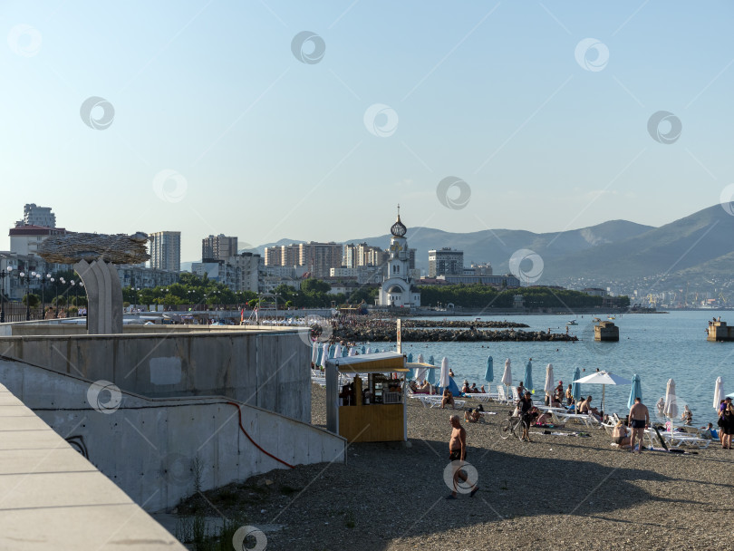 Скачать 12 августа 2025 года: Городской пляж в Новороссийске, Россия, рядом с памятником Хамсе, на заднем плане видны церковь и горы. фотосток Ozero