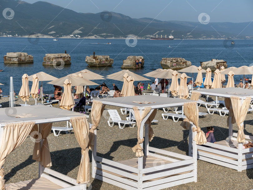 Скачать 12 августа 2025 года: Люди отдыхают на городском пляже в Новороссийске, Россия, в то время как вдалеке по Черному морю плывет грузовой корабль. фотосток Ozero