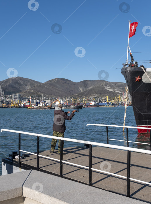 Скачать 22 августа 2025 года рыбак забрасывает удочку с пирса возле авианосца "Адмирал Кузнецов" в Новороссийске, Россия, на побережье Черного моря. фотосток Ozero