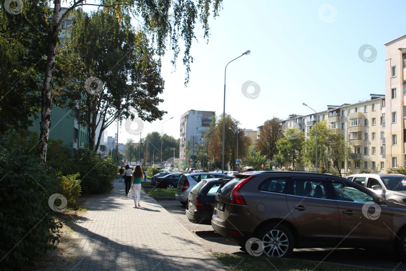 Скачать Беларусь, Минск - 22.09.2024: Тихий городской тротуар с пешеходами. Солнечный свет освещает оживленную городскую улицу с припаркованными автомобилями фотосток Ozero