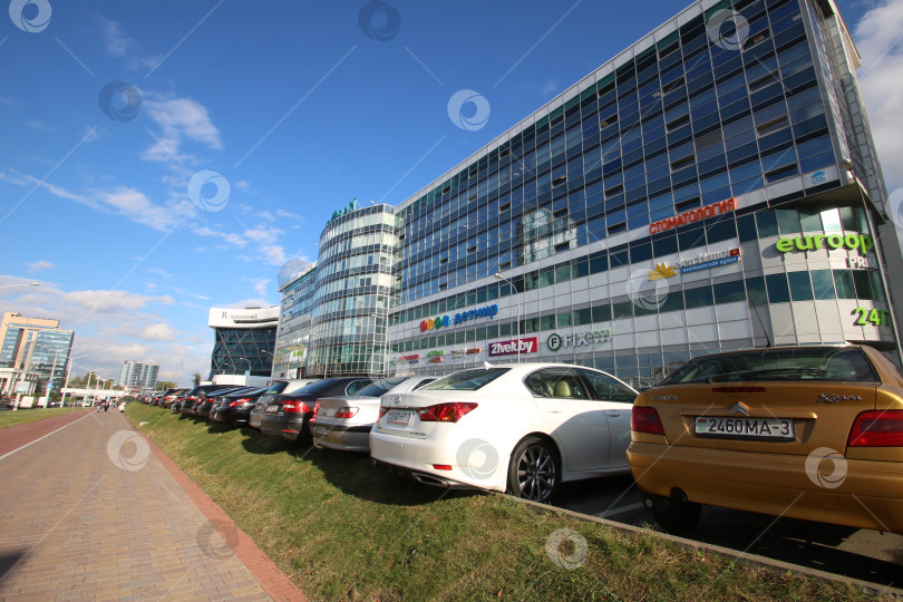 Скачать Беларусь, Минск - 22.09.2024: Городской пейзаж с отражениями. Современная городская сцена с блестящими стеклянными фасадами и припаркованными автомобилями фотосток Ozero
